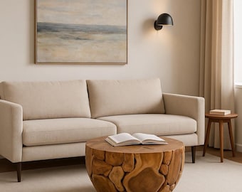 Op de afbeelding: Een woonkamer met een beige bank, een ronde houten salontafel met een getextureerd ontwerp en een ingelijst landschapsschilderij. Een kleine houten bijzettafel en een zwarte wandlamp zijn ook zichtbaar. Neutrale sfeer.