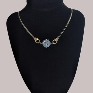 Puede incluir: Un collar de cadena en tono dorado que presenta un grupo central de cuentas facetadas de color azul claro. La cadena está conectada al grupo central de cuentas mediante dos pequeños dijes de metal dorado con forma de esposas.