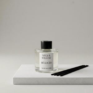 May include: A clear glass diffuser bottle with a black cap and a label that reads "WICK & WILLOW BELLAGIO". The bottle sits on a white square platform with black reeds next to it. The net weight is 4oz (118ml).