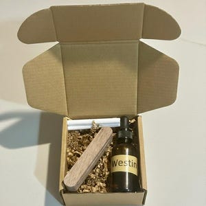 May include: A brown cardboard box with an open lid, containing a brown glass bottle with a dropper, a wooden block, and white sticks. The bottle has a white label with the word "Westin" printed on it. The box is filled with brown packing material.