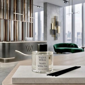 May include: A clear glass bottle of Wick & Willow Westin White Tea fragrance with a black cap, sitting on a white marble surface. The bottle is labeled with the product name and net weight of 4 oz (118 ml). Black reed sticks are next to the bottle.