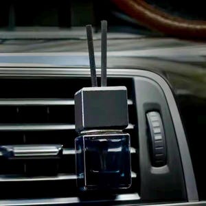 May include: A black car air freshener with a square top and a rectangular glass bottle. Two black reeds protrude from the top. The air freshener is hanging from a car vent.