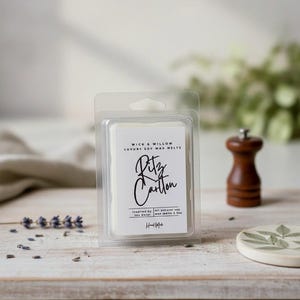 May include: A clear plastic package containing white soy wax melts. The label reads "Ritz Carlton" in elegant script, with additional text indicating the product is inspired by the hotel. A wooden pepper grinder and a small ceramic coaster with a leaf design are also visible.