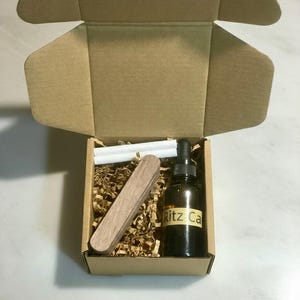 May include: An open cardboard box containing a brown glass bottle with a dropper, a wooden tool, and two white sticks, all nestled in brown packing material. The bottle has a label that reads "Ritz Ca".