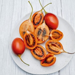 Könnte beinhalten: Auf einem weißen, gesprenkelten Teller liegen mehrere Tamarillo-Früchte, einige ganz und andere halbiert. Die ganzen Früchte sind leuchtend rot, während die geschnittenen ein dunkles Inneres und orangefarbenes Fruchtfleisch zeigen. Grüne Stiele sind sichtbar.