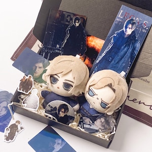 May include: A black gift box containing two plush dolls with blonde hair and blue eyes, alongside various printed cards, stickers, and acrylic charms featuring a male character. The items include a tall bookmark labeled Leon S. Kennedy.