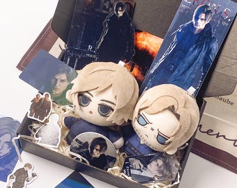 May include: A black gift box containing two plush dolls with blonde hair and blue eyes, alongside various printed cards, stickers, and acrylic charms featuring a male character. The items include a tall bookmark labeled Leon S. Kennedy.
