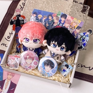 May include: A gift box containing two plush dolls with pink and black hair, two circular pin-back buttons, two acrylic character standees, and two square acrylic keychains featuring anime-style illustrations of characters with various expressions.
