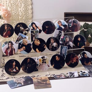 May include: A collection of circular pin-back buttons featuring various character portraits and illustrations from the Final Fantasy VII video game series, displayed on a clear tiered acrylic stand alongside several rectangular trading cards.