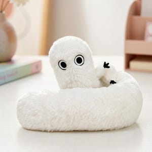 May include: A white plush toy shaped like a long, coiled worm with a soft, fuzzy texture. It features two large, circular eyes with black pupils and white irises, along with small, black, three-fingered hands extending from its body.