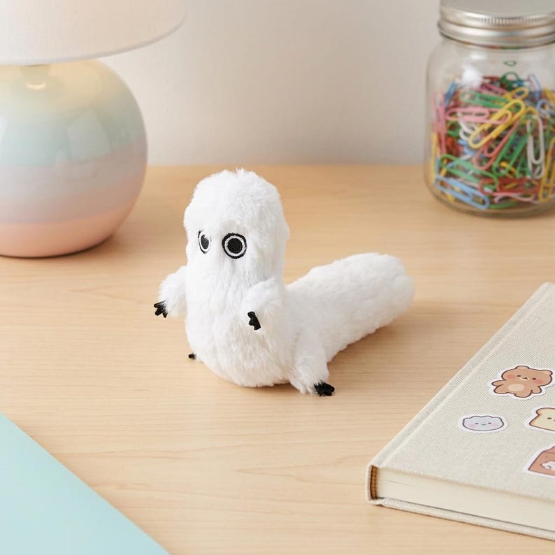 May include: A small, white, plush creature with soft fur, two large circular black eyes, and tiny black-tipped limbs. The figure has an upright head and a long, horizontal body extending to the right, featuring a simple, rounded design.