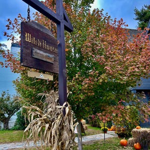 May include: A dark brown wooden signpost hanging a rectangular wooden sign that reads Witch House Salem Mass. The post is decorated with dried corn stalks at the base and features a small black sign at the bottom that reads Please Keep Off The Grass.