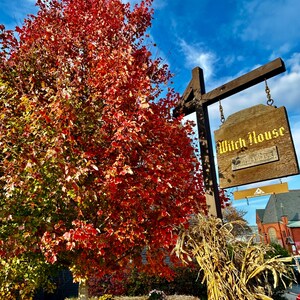 May include: A wooden sign for the Witch House hangs from a brown post, with a backdrop of a vibrant red tree and a bright blue sky. Pumpkins and hay bales are at the base of the tree, with dried corn stalks to the right.