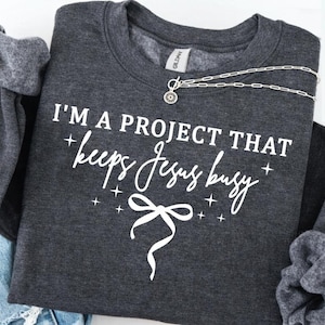 May include: Dark gray sweatshirt with the text "I'M A PROJECT THAT keeps Jesus busy" in white script. A silver necklace with a small pendant is draped across the front. The sweatshirt is folded, with the sleeves visible.