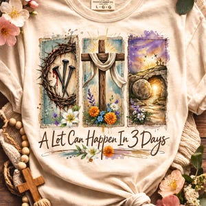 May include: Cream-colored t-shirt featuring a watercolor design with religious imagery. The design includes a crown of thorns, a wooden cross, and an empty tomb, with the text "A Lot Can Happen In 3 Days".