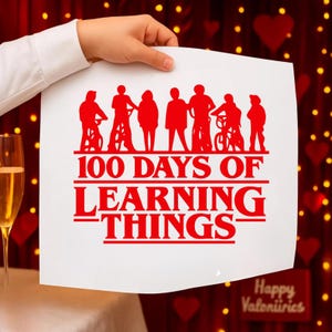 以下が含まれることがあります： 赤い文字とグラフィックが描かれた白いポスターが手で持たれています。テキストは「100 DAYS OF LEARNING THINGS」と書かれており、テキストの上には人々と自転車のシルエットがあります。背景にはシャンパングラスとバレンタインデーの装飾が含まれています。
