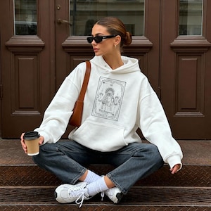 Può includere: Una felpa con cappuccio bianca con una grafica nera di una carta dei tarocchi, "The Empress", indossata con jeans scuri e scarpe da ginnastica bianche. Una borsa a tracolla marrone e occhiali da sole completano il look. Viene tenuta una tazza di caffè.