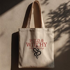 Può includere: Borsa tote in tela beige con le parole "WILD & WITCHY Stay Whimsy" in rosso e nero. Un disegno di serpente e floreale nero è sotto il testo. La borsa ha due manici beige.