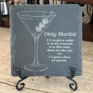 May include: A square dark gray slate coaster featuring an etched illustration of a martini glass with three olives on a pick. The text reads: Dirty Martini, 2 1/2 oz gin or vodka, 1/2 oz dry vermouth, 1/2 oz olive brine, Ice, 2-3 green olives.