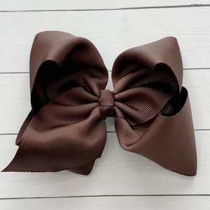 May include: A large, brown hair bow made of grosgrain ribbon. The bow is a rich, chocolate brown color and has a classic, oversized design. The ribbon is textured with vertical ridges.