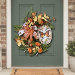 May include: A decorative wreath featuring orange and yellow artificial flowers, green fern foliage, and several butterfly accents. An oval sign with a monarch butterfly illustration and floral pattern is attached, along with a plaid orange ribbon bow.