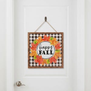Könnte beinhalten: Ein quadratisches Wandbild aus Holz mit einem schwarz-weißen Karomuster im Hintergrund. Ein kreisförmiges Zentrum enthält den Text "happy FALL" in schwarzer Schrift, umgeben von einem Kranz aus orangefarbenen, roten und grünen Ahornblättern.