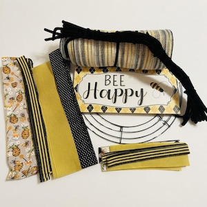 May include: A craft kit featuring a rectangular sign with the text BEE Happy, a round wire wreath frame, a roll of yellow and black mesh, black pipe cleaners, and several fabric ribbons in yellow, black polka dot, and bee-patterned prints.
