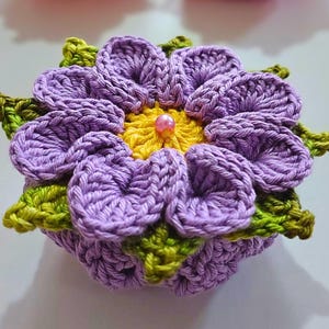 Könnte beinhalten: Eine handgefertigte Häkelblume in Lila, Gelb und Grün. Die Blume hat mehrere Schichten von Blütenblättern und eine kleine Perle in der Mitte. Weitere Häkelblumen in Orange und Rot sind im Hintergrund zu sehen.