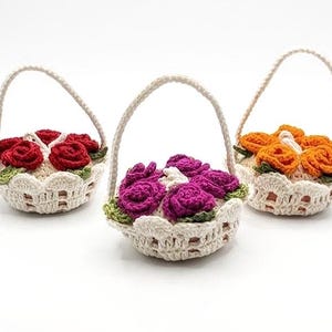 Pode incluir: Três cestos de crochê feitos à mão, cada um cheio de rosas coloridas. Os cestos têm uma base creme com uma borda ondulada e uma alça a condizer. Um cesto contém rosas vermelhas, outro roxas e o terceiro laranja.