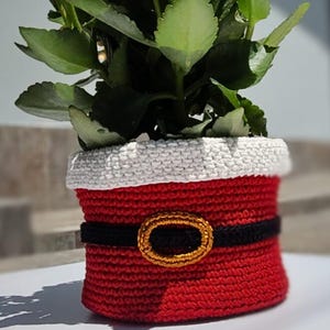 Puede incluir: Una maceta roja de ganchillo con ribete blanco, cinturón negro y hebilla dorada, que se asemeja al traje de Papá Noel. Contiene una planta floreciente con hojas verdes y flores naranjas, una decoración festiva.