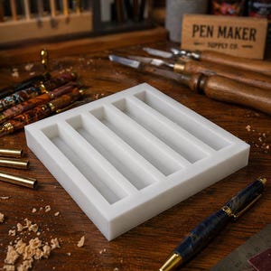 May include: A white rectangular pen holder with five slots sits on a wooden surface. A finished pen with a blue and gold design rests nearby. The background includes tools, pens, and a sign that reads "PEN MAKER SUPPLY CO."