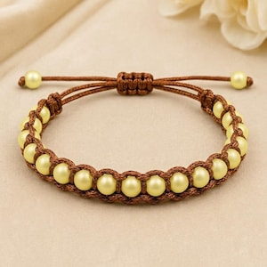 Può includere: Un braccialetto caratterizzato da una serie di perline sferiche giallo pallido intrecciate in un cordino marrone. Il design include una chiusura a nodo scorsoio regolabile con due perline gialle più piccole alle estremità del cordino.