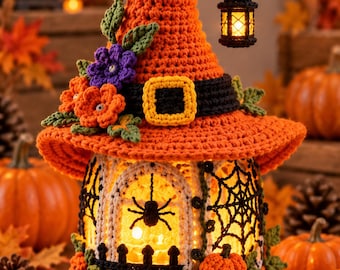 May include: A crocheted orange witch hat topper sits atop a cylindrical structure featuring a black spider web design, a small black fence, and a glowing interior. The hat is adorned with purple and orange flowers and a black band with a yellow buckle.