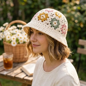 Może przedstawiać: Szydełkowy kapelusz typu bucket hat z kremową bazą i powtarzającym się wzorem wielokolorowych motywów kwiatowych w odcieniach musztardowej żółci, szałwiowej zieleni i delikatnego różu. Kapelusz ma zaokrągloną główkę i krótkie, opadające rondo.