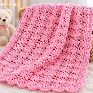 May include: A bright pink crochet baby blanket featuring a repeating fan-like stitch pattern and a scalloped edge. The blanket is draped over a woven wicker basket, showcasing the intricate openwork texture of the yarn throughout the entire piece.