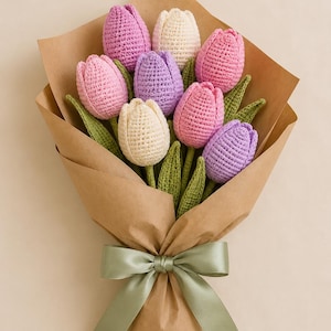 Op de afbeelding: Een boeket van gehaakte tulpen in tinten lichtroze, lavendel en crème, voorzien van groene gehaakte bladeren. De bloemen zijn verpakt in bruin papier en vastgebonden met een saliegroen satijnen lint, wat een gestructureerd bloemstuk vormt.