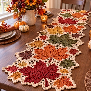 Può includere: Un lungo runner da tavola rettangolare all'uncinetto con un motivo ripetuto di foglie d'acero nei toni del rosso, arancione, giallo, marrone e verde, unito da un bordo in pizzo color crema che crea un profilo smerlato lungo tutta la lunghezza.