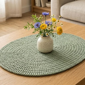 Può includere: Una tovaglietta ovale all'uncinetto color verde salvia con un motivo a maglia testurizzato. Al centro della tovaglietta si trova un piccolo vaso in ceramica bianco sporco maculato contenente un bouquet di ranuncoli gialli e margherite.