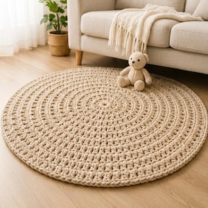 May include: A large, circular, beige crochet rug featuring a thick, textured yarn construction with a repeating concentric pattern. The rug is crafted from chunky cord-like material, creating a dense, soft surface with a consistent, neutral colour.