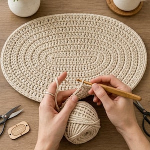 Pode incluir: Um tapete oval de crochê bege feito de cordão grosso, um novelo de fio bege correspondente, uma agulha de crochê de madeira, uma tesoura preta e uma pequena etiqueta retangular de madeira com um padrão oval gravado sobre madeira.