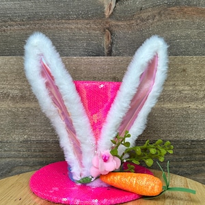 May include: A pink sequined top hat with attached white fluffy bunny ears. The hat is decorated with a pink flower, green sprigs, and an orange carrot. The hat is designed for Easter.