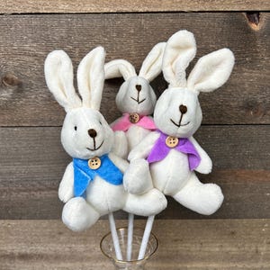 May include: Three white plush bunny decorations with long ears, each wearing a different colored bow tie: blue, pink, and purple. The bunnies are attached to white sticks and placed in a clear glass vase with a gold rim. The background is a wooden wall.