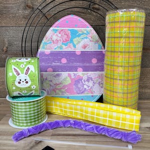 May include: An assortment of Easter craft supplies, including a decorative egg, rolls of ribbon, and a roll of yellow plaid mesh. The egg features floral and polka dot patterns. A green ribbon with a bunny design is also visible.