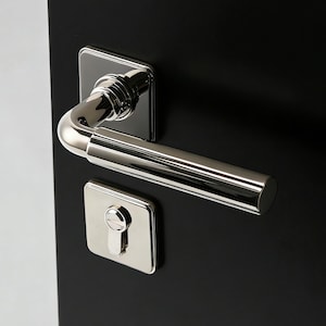 Peut inclure: Une poignée de porte et une plaque de trou de serrure en chrome poli sont montées sur une porte noire. La poignée est de style levier incurvé, et la plaque de trou de serrure est carrée. La porte est noire unie.
