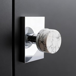 May include: A modern door knob with a polished chrome square backplate and a cylindrical knob made of white marble with gray veining. The knob is mounted on a dark gray door, showcasing a minimalist design.