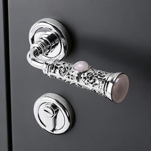Puede incluir: Un juego de manija de puerta cromada pulida con un diseño calado decorativo. La manija presenta un acento de piedra preciosa rosa claro y una cerradura de puerta cromada a juego. La ferretería está colocada sobre una puerta gris oscuro.