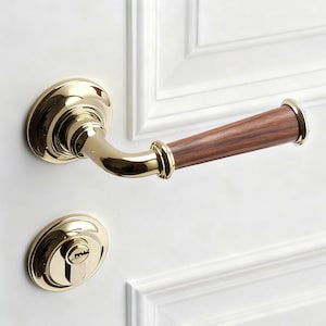 May include: A polished gold door handle and keyhole escutcheon with a dark wood handle. The handle is curved and has a classic design, mounted on a white door with paneling.