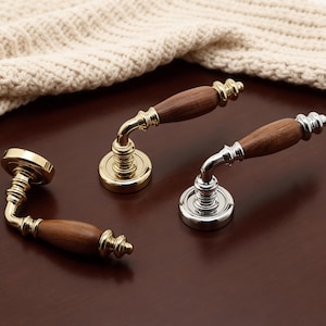 Puede incluir: Tres manijas de puerta con empuñaduras de madera y bases metálicas. Dos son doradas y una plateada. Las manijas son curvas con un diseño decorativo donde la empuñadura se encuentra con la base. Sobre una superficie marrón oscuro.