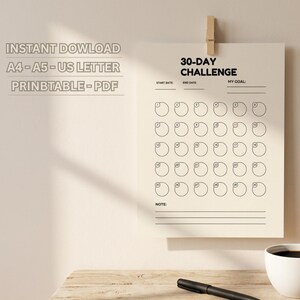 Puede incluir: Un imprimible beige de desafío de 30 días, sujeto con una pinza de madera. El texto "30-DAY CHALLENGE" está en la parte superior, con espacios para las fechas de inicio y fin, y un objetivo. Debajo hay 30 círculos para el seguimiento. Una taza de café blanca, un bolígrafo y una pequeña planta en maceta están sobre la mesa.