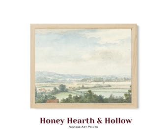 May include: A landscape art print in a light wood frame featuring a watercolor painting of a rural valley with rolling hills, green fields, and scattered houses under a pale blue sky. The text Honey Hearth & Hollow Vintage Art Prints is below.